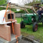 Ein Lisboa Holzbackofen wird mit einem Traktor und Frontlader auf ein Gestell gehoben